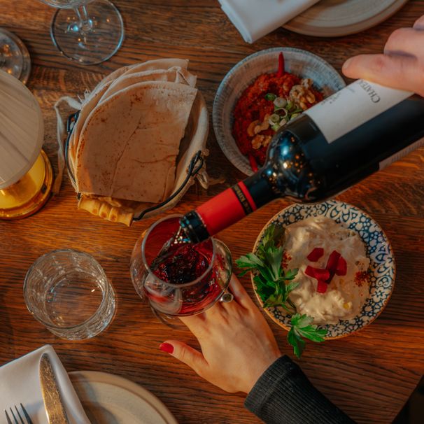Rött vin hälls upp vid ett bord med hummus och bröd, sett ovanifrån